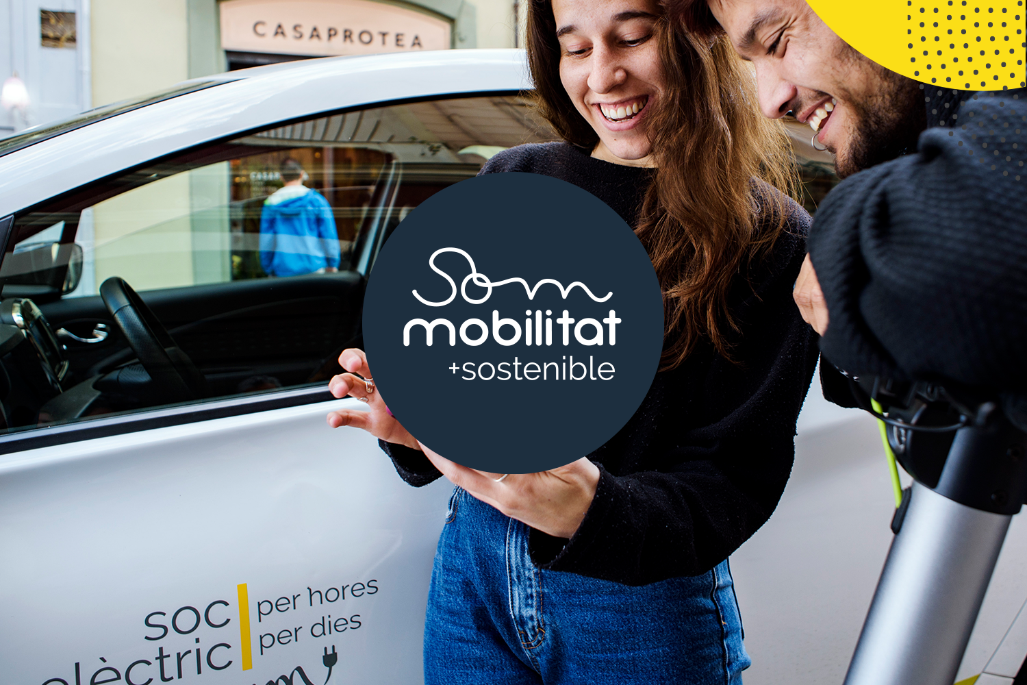 Image with to persons in front of a car. One person which can be read as female holds a smartphone in her hand and shows the display to the other person. The car is an electric car which belongs to the company of Som Mobilitat, an electric carsharing cooperative.
